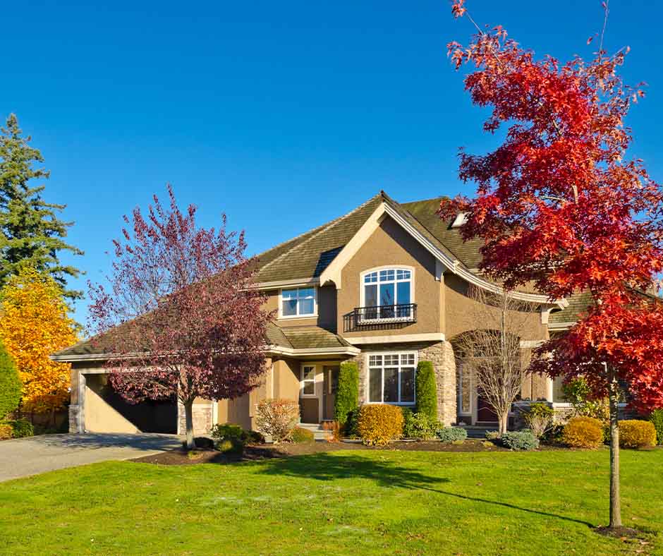 Two-story suburban house with a landscaped front yard and colorful autumn trees under a clear blue sky.